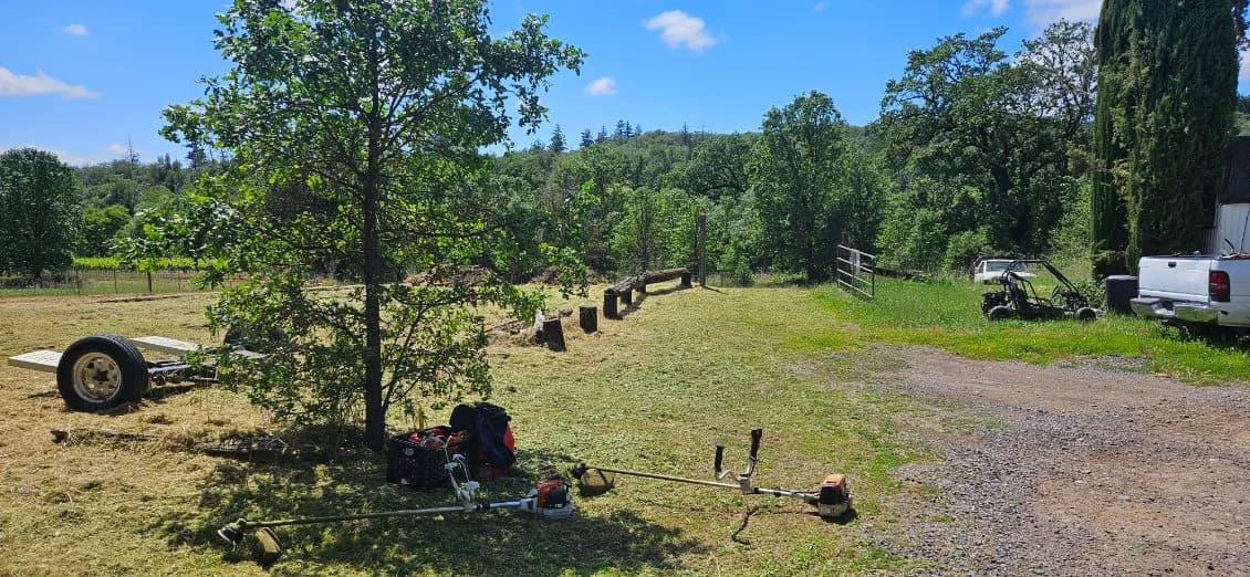 Maples Landscaping field-clearing equipment laid out under a tree — multiple string trimmers, a UTV, and a work truck on a Mendocino County rural property